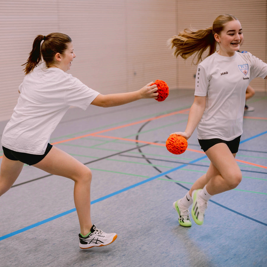 Zwei Mädchen spielen in der Sporthalle Fangen, beide halten einen orangefarbenen Ball